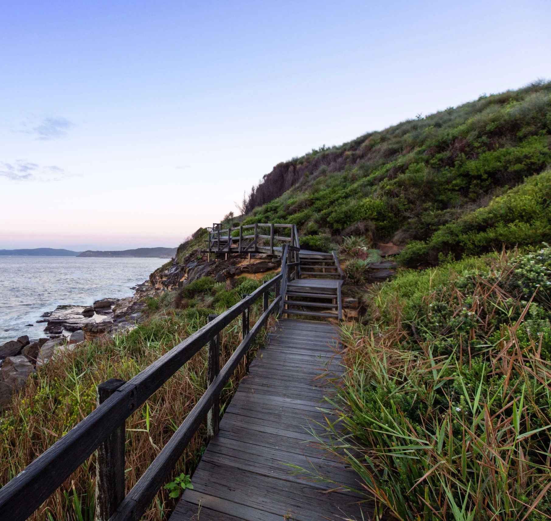 Wooden pathway along the rocky coastline with green bushes and hillside, overlooking a body of water under a clear sky.