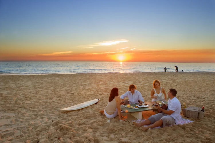 A couple enjoys a romantic sunset picnic on a beach in the Whitsundays, Queensland, Australia, with golden light reflecting off the calm sea and soft white sand.