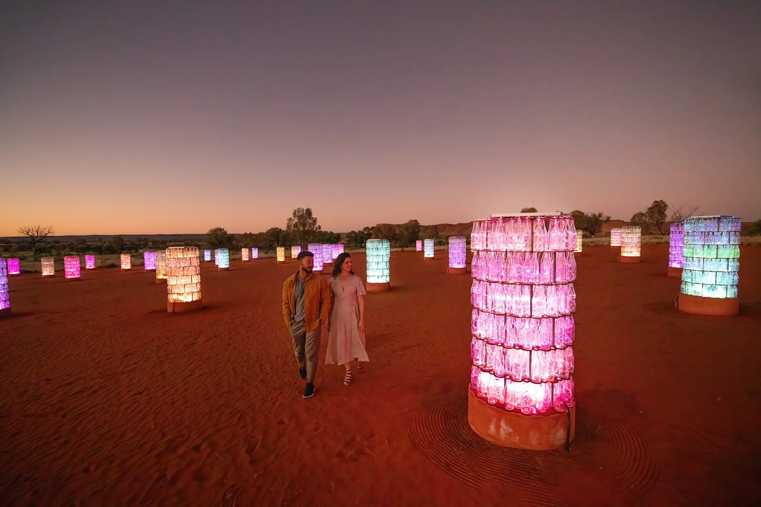 Private viewing experience with a couple walking through the Light-Towers installation in Kings Canyon, Red Centre, Australia, featuring glowing glass towers illuminated against the desert landscape at dusk.