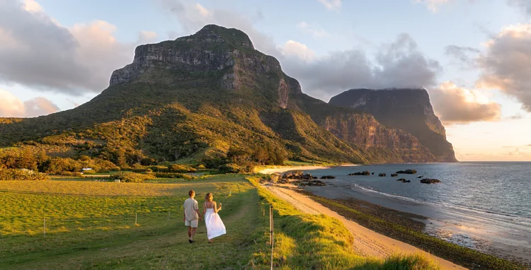 A couple walking along a secluded beach on the Seven Peaks Walk at Lord Howe Island, New South Wales, Australia, with turquoise waves lapping at the shore and lush green peaks rising in the background.