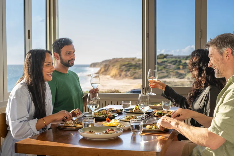 A family shares a wine-country lunch at a table overlooking the coast, raising glasses together over a spread of food.
