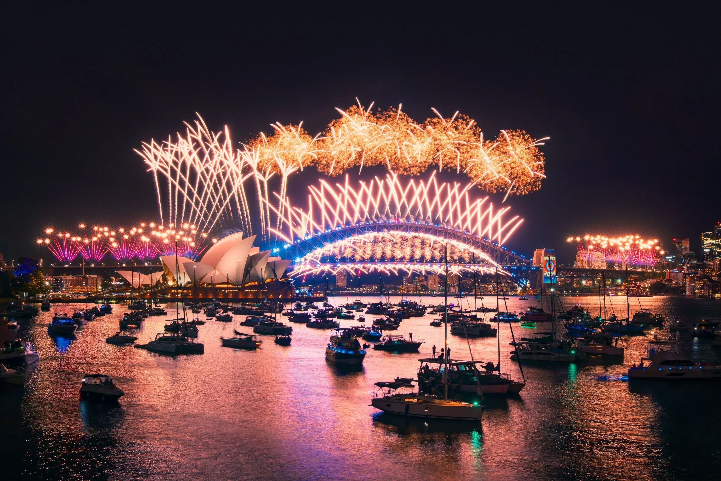Nighttime fireworks over the Sydney Harbour Bridge and Sydney Opera House with boats on the water.