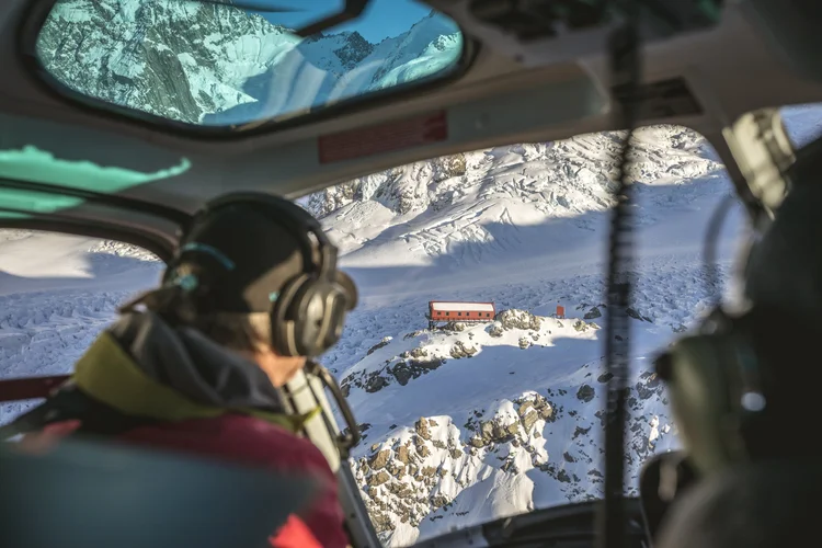 View from inside a ski plane cockpit flying over a snow-covered glacier and mountains, with a small red hut visible on the ice below.