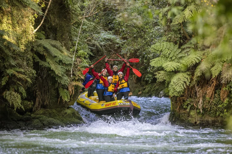 A family in helmets and life jackets paddles a yellow raft through whitewater rapids on a forest-lined river.