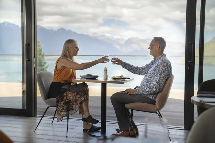 An older couple toasting wine glasses at an outdoor table overlooking a lake and mountains in New Zealand