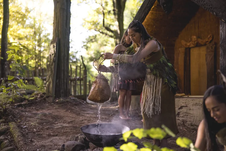 Two people in traditional Māori dress prepare a hāngī, lowering food into a steaming earth oven in front of a carved wharenui.