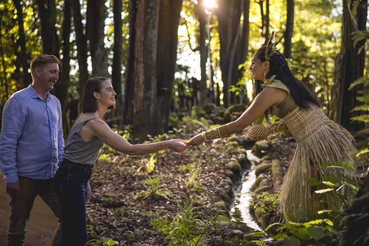A Māori woman in traditional dress greets a visitor with a handshake in a native forest clearing.