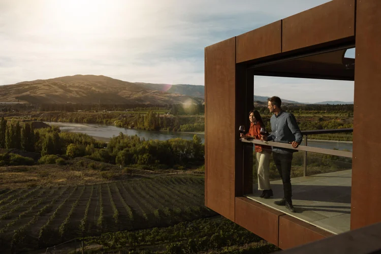 A couple standing on the balcony of a modern lodge overlooking a river valley and mountains.