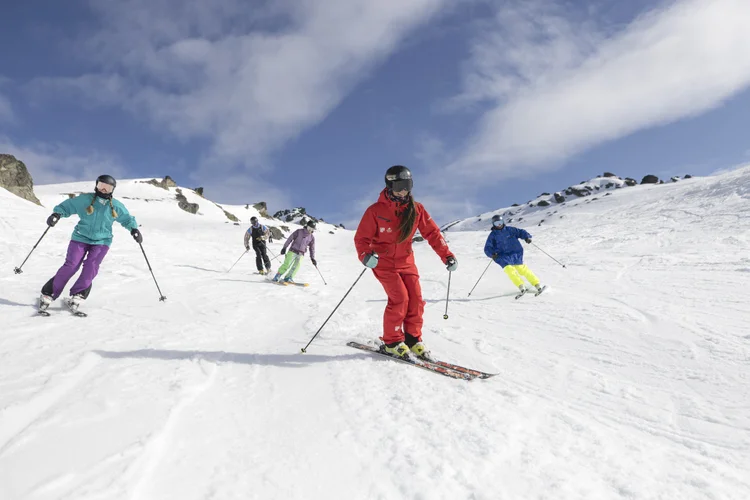 A skier in a red suit leads a small group of skiers down a snowy alpine slope under a clear blue sky.