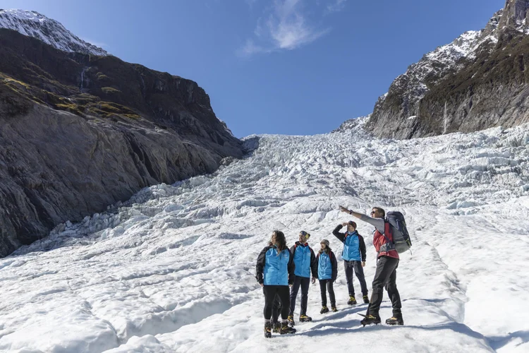 A guide leads a group of helmeted hikers, including children, across a glacier beneath steep mountain cliffs.