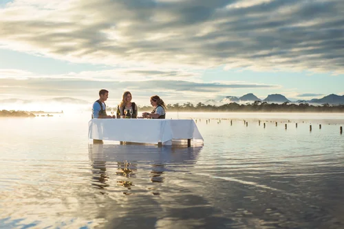 A couple and a woman having a dinner on a floating table in a body of water during sunset with mountains and trees in the background.