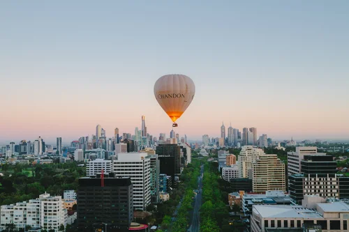 A hot air balloon floating above a city skyline at sunset, with tall buildings and a tree-lined street below.