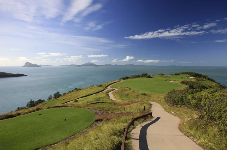 A coastal golf course with lush green fairways, walking path, and a view of the ocean and distant islands under a partly cloudy sky.