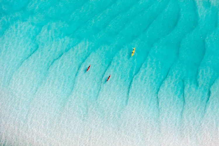 Aerial view of three kayakers paddling in clear, turquoise water with visible dark shadows beneath the surface.