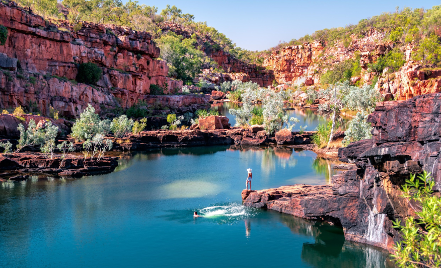 A person standing on a rock ledge overlooking a river in a canyon, with one person swimming in the water. The canyon has red rock walls and is dotted with green trees, under a clear blue sky.