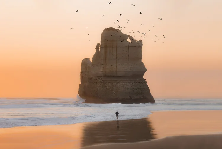 Person walking on the beach at sunset with a large rock formation and birds flying overhead.