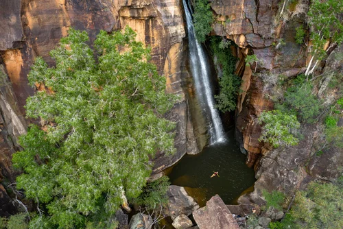 An aerial view of a small waterfall flowing down a red-brown rocky cliff into a dark pool of water, surrounded by green trees and rocks.