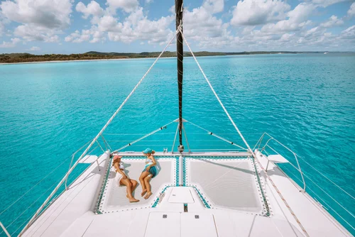 Two women in swimsuits lying on a trampoline on a sailboat in clear turquoise water, with an island and blue sky with clouds in the background.