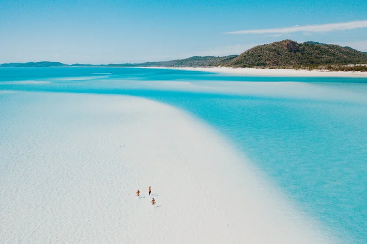 Aerial view of a pristine white sand beach with turquoise waters and distant green hills under a blue sky.