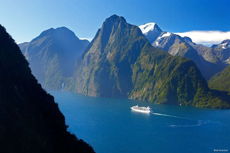 A large cruise ship sailing in a deep blue fjord surrounded by steep, green mountains with some snow on their peaks.