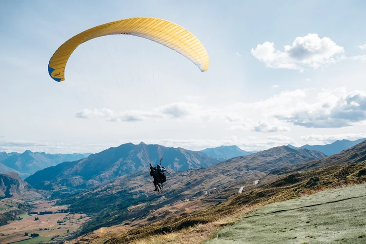 Two people paragliding over a mountain valley with grassy hills and distant mountain peaks under a partly cloudy sky.