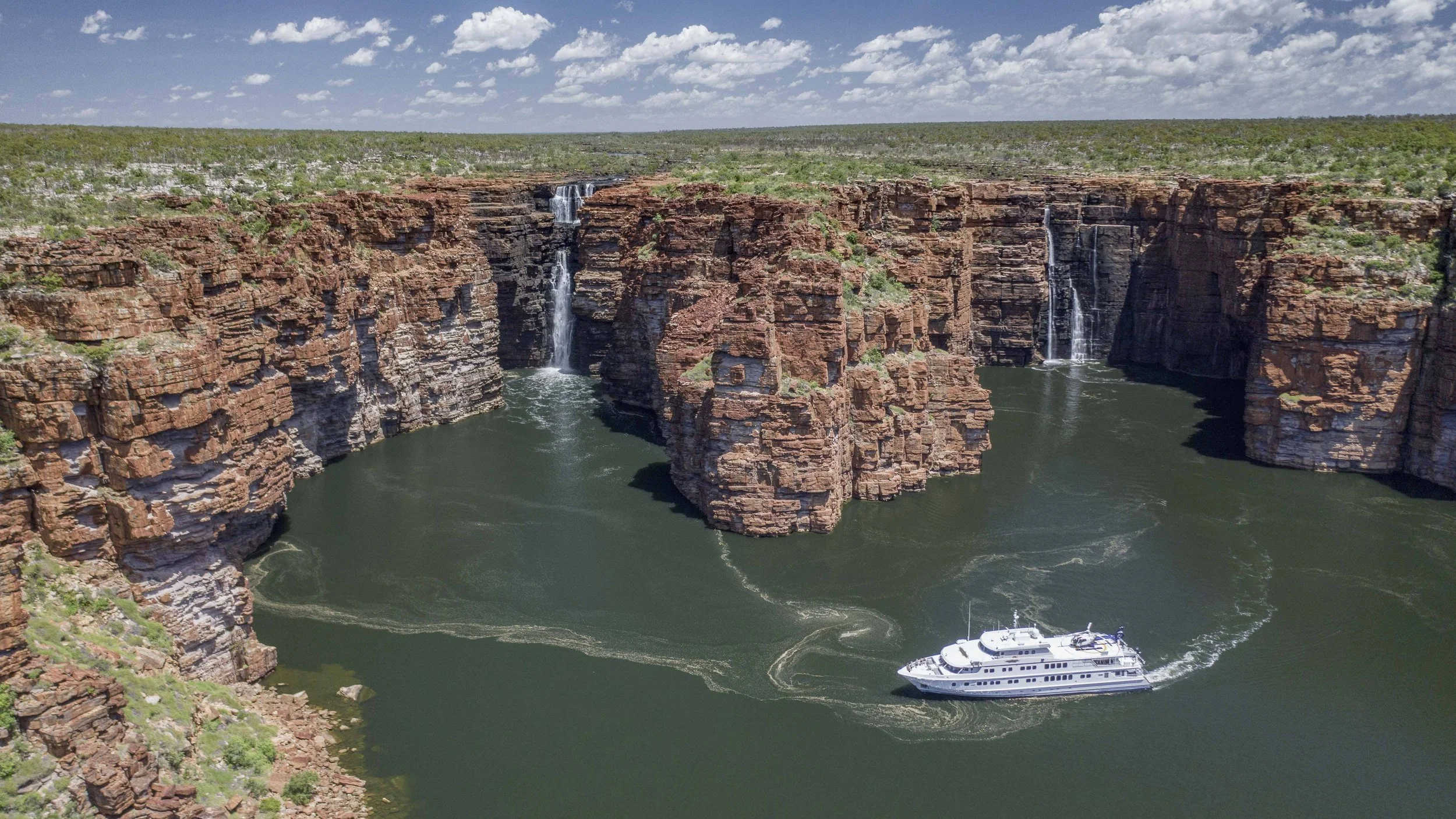 Aerial view of a large canyon with waterfalls flowing into a river, a white yacht sailing in the water, and a clear sky with scattered clouds.