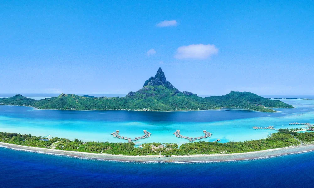 Aerial view of a tropical island with a mountain peak, surrounded by a lagoon and coral reefs, with overwater bungalows on the water and a lush green shoreline