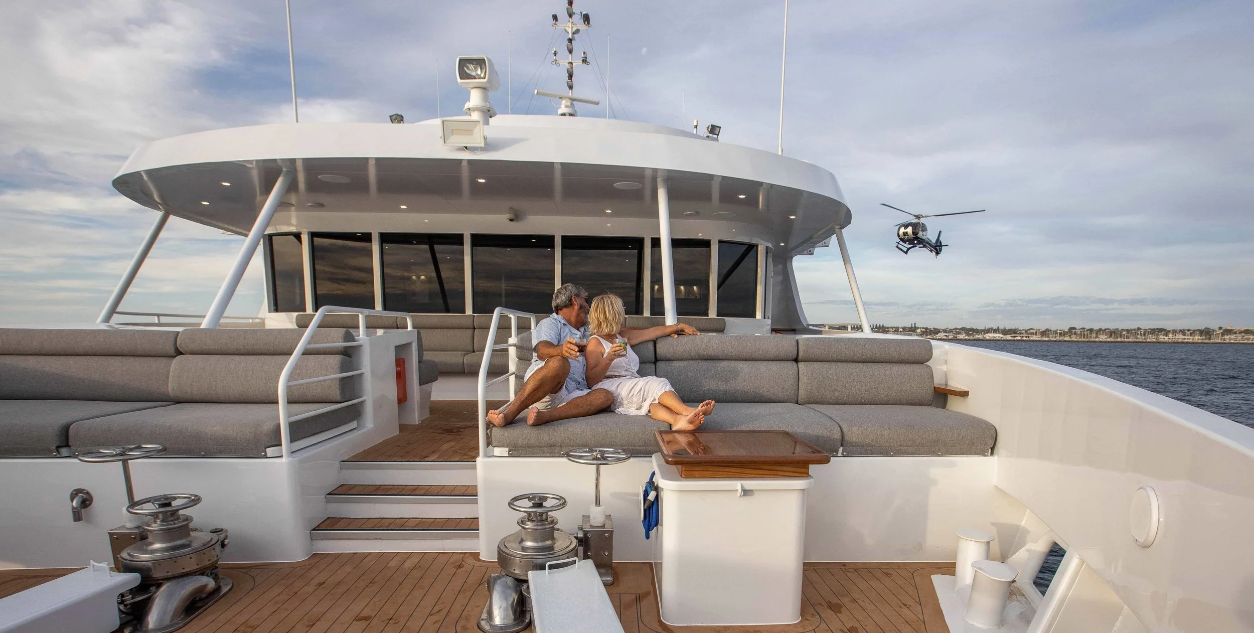 Couple sitting on the deck of a luxury yacht, with a helicopter flying nearby over water.