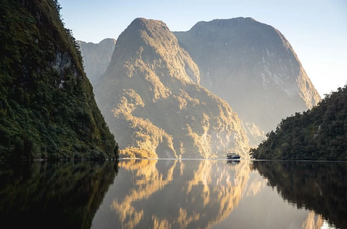 A boat sailing on a calm river surrounded by towering green mountains with reflected images on the water.