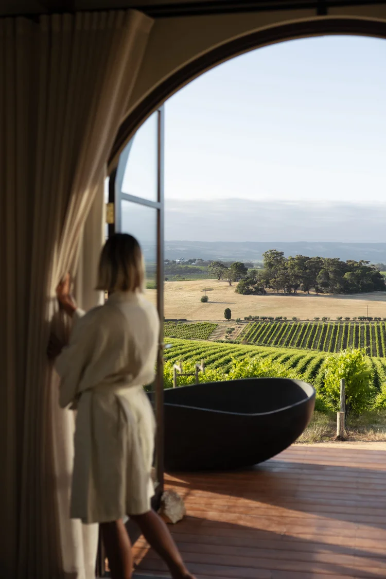 A person stands in a white robe beside an outdoor bath, ready to enjoy the idyllic view from their accommodation nestled in the heart of a vineyard in McLaren Vale, South Australia.