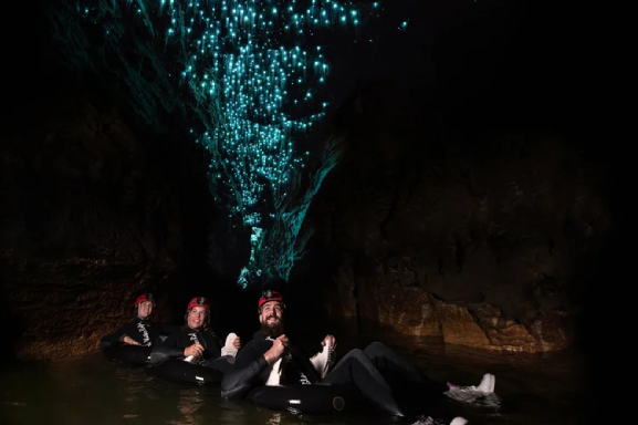 Family floats on inner tubes through a dark underground cave lit by thousands of glowing blue-green glowworms overhead.
