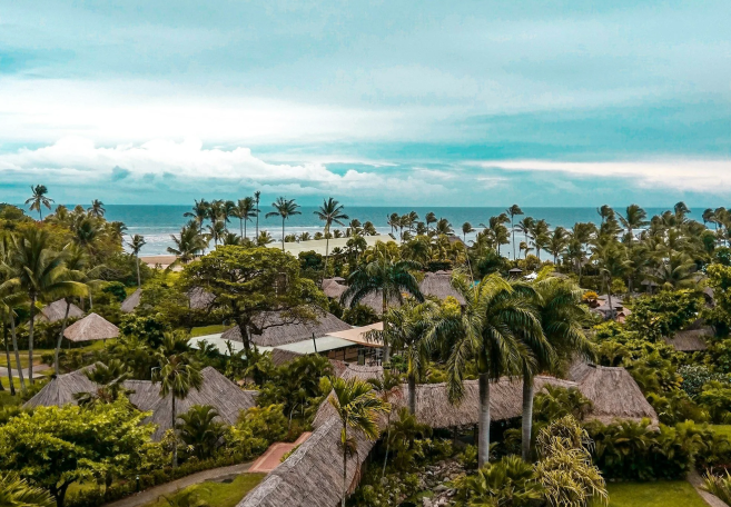 A tropical resort with thatched roof buildings, surrounded by lush green trees and plants, near a beach with palm trees and ocean in the background under cloudy sky.