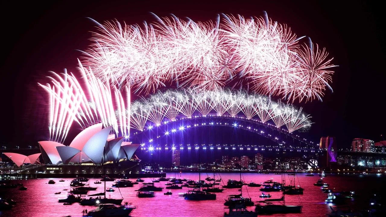 Fireworks display above the Sydney Opera House and Sydney Harbour Bridge at night, with boats on the water reflecting colorful lights.