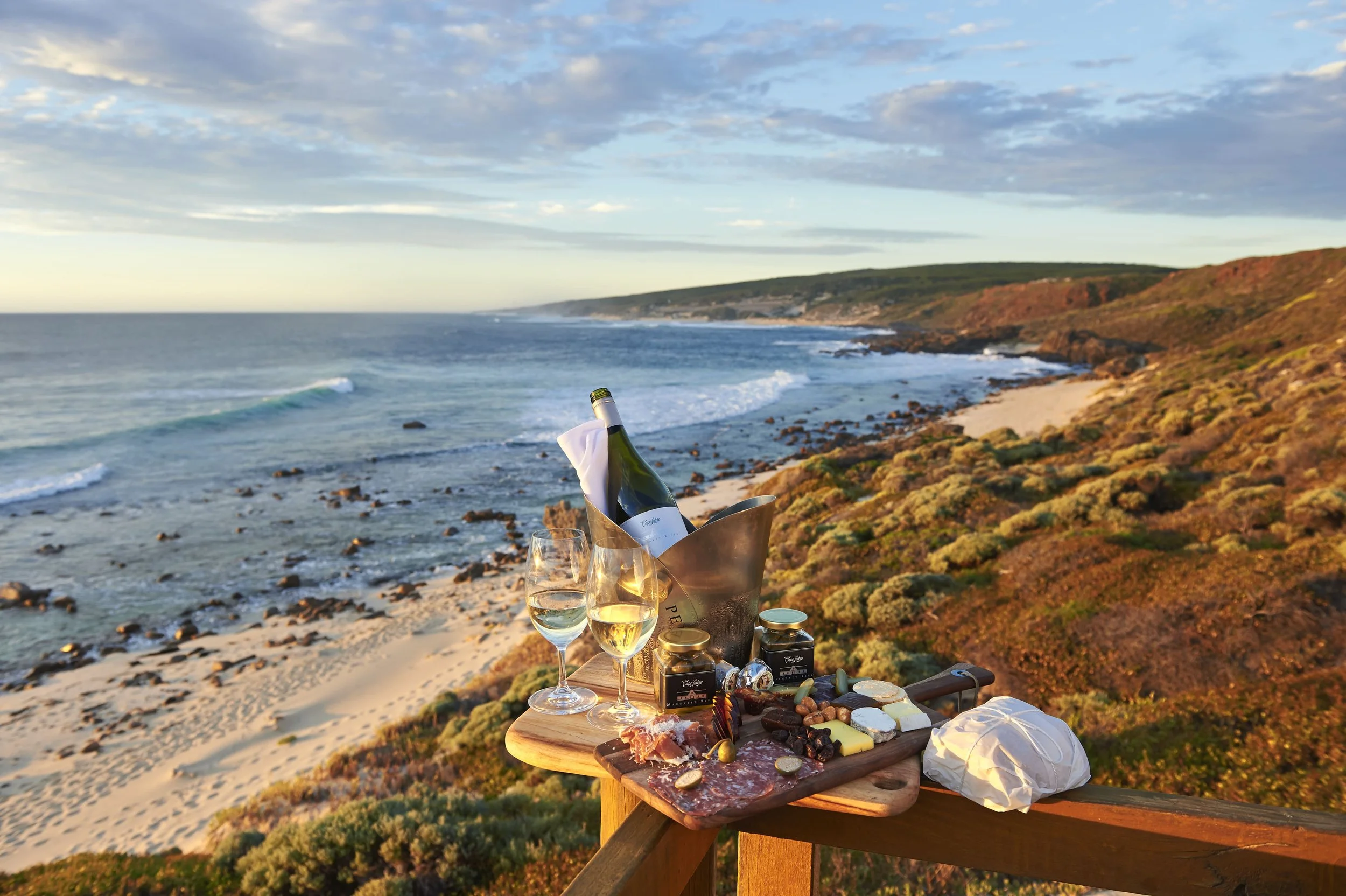 A picnic setup on a wooden railing overlooking a beach with rocks and gentle waves, featuring a bottle of white wine in an ice bucket, two wine glasses filled with white wine, a cheese platter with various cheeses, meats, and accompaniments, and small jars of jam or preserves, with a scenic coastal backdrop under a partly cloudy sky in the evening.