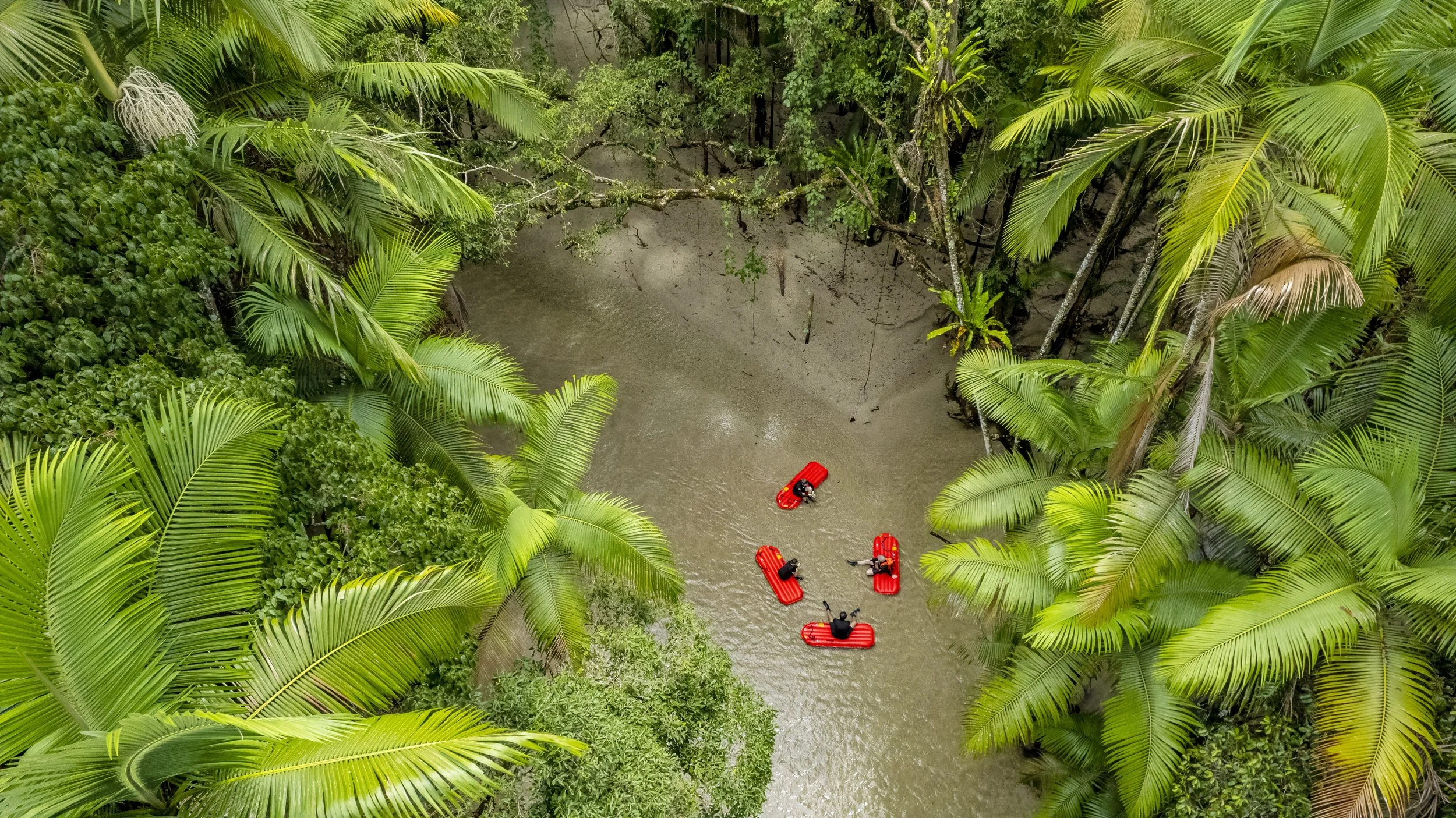 Five people with life jackets and helmets rafting on a river surrounded by lush green tropical vegetation.