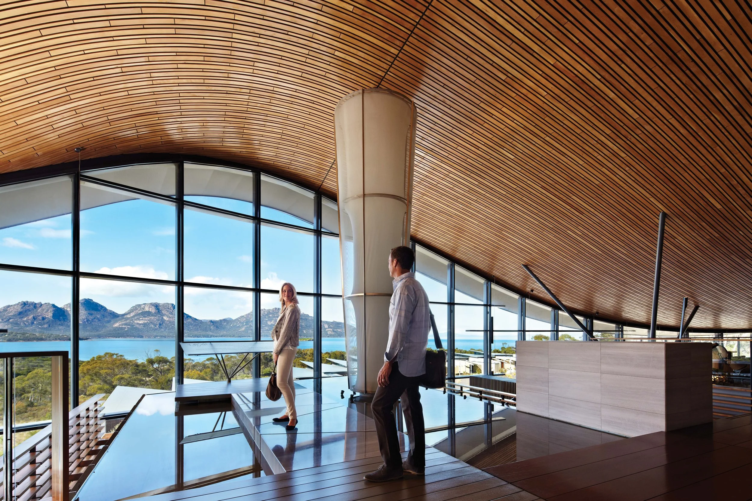 Interior of a modern building with large glass windows overlooking mountains, lake, and trees, with two people standing inside - a woman looking outside and a man walking with a briefcase.