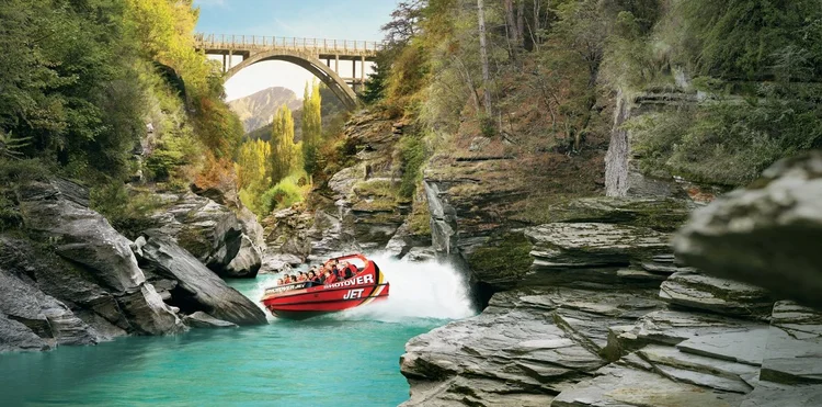 A red jet boat speeding through a narrow rocky canyon with turquoise water, trees on either side, and a stone bridge overhead.