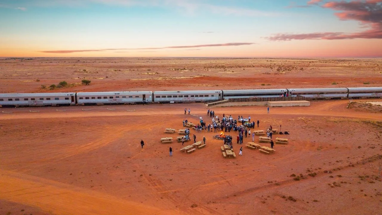 People gathered around a campfire with benches in a desert, a train passing by in the background at sunset.