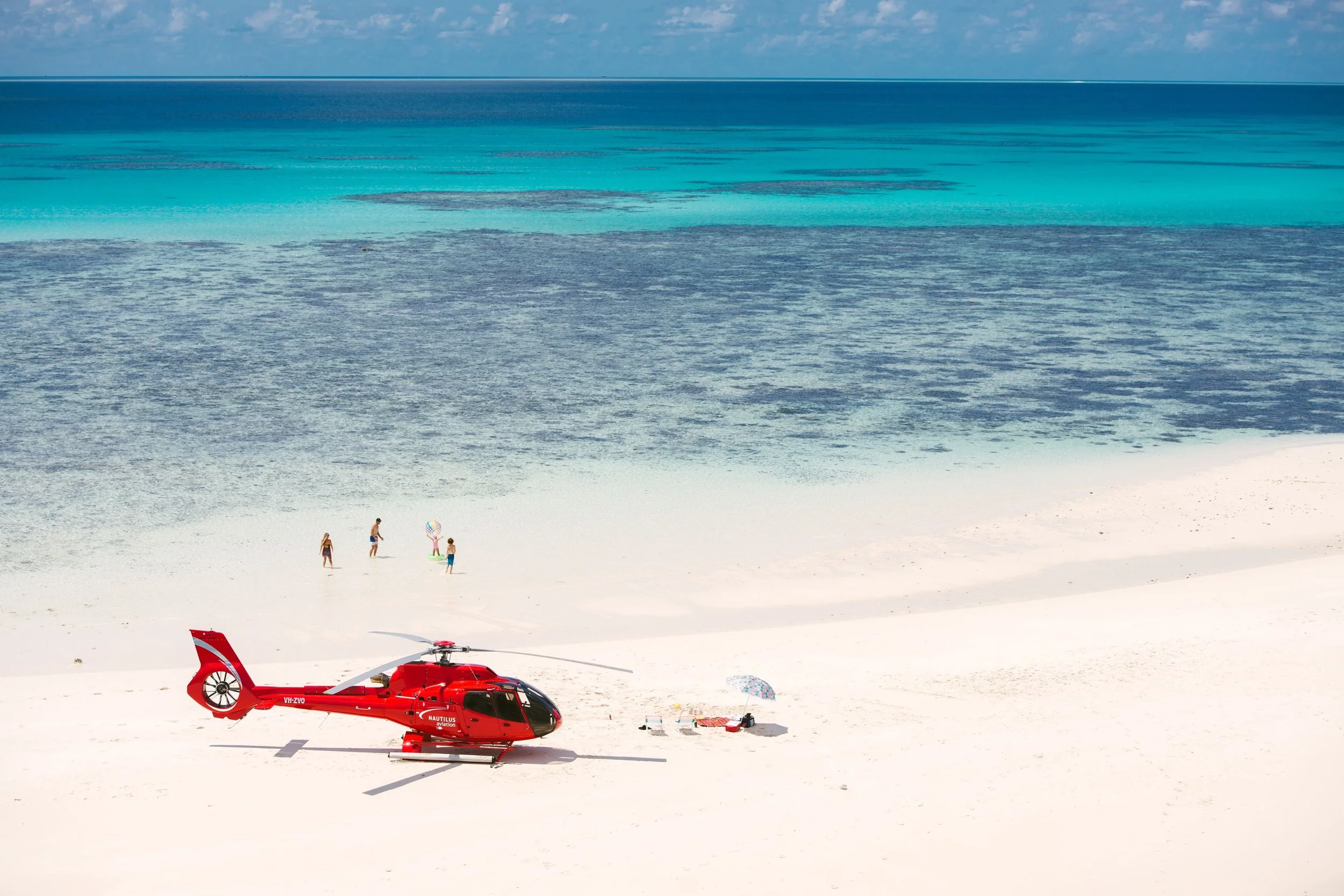 A red helicopter on a white sandy beach near the ocean, with a few people in the distance, some under umbrellas and holding beach toys, in a tropical setting.