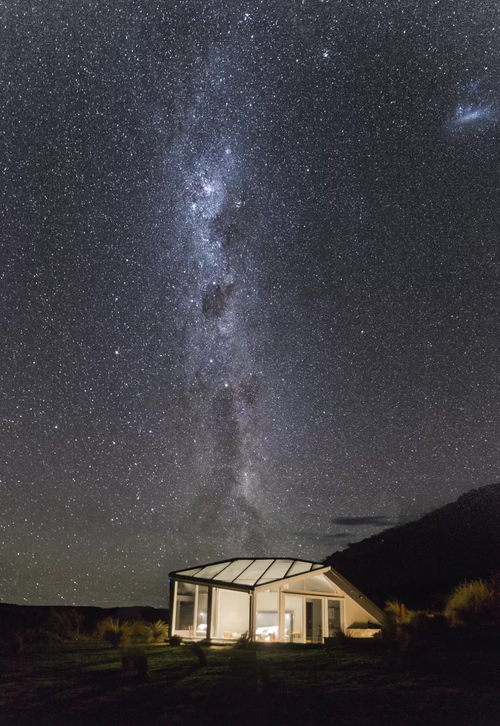 A house with large glass windows and a sloped roof under a starry night sky with the Milky Way galaxy visible.