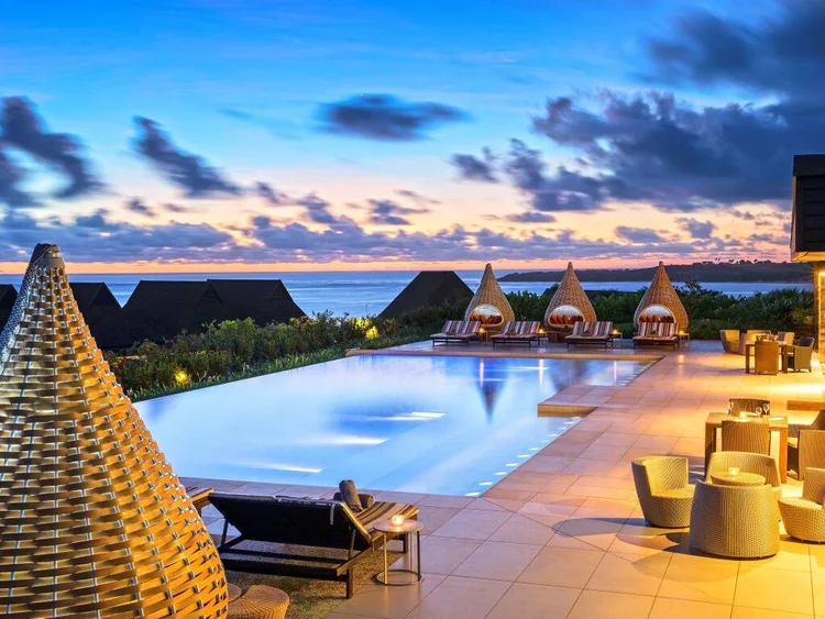 Infinity pool at dusk with a pastel sunset sky, thatched-roof cabanas, and wicker lounge furniture in the foreground.
