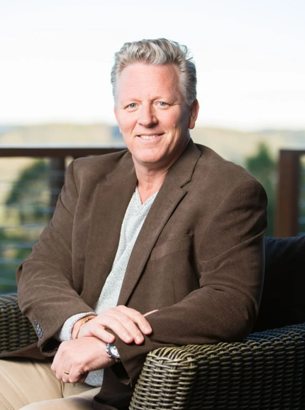 A middle-aged man with gray hair, casually dressed in a brown blazer and beige pants, sitting on a wicker outdoor chair with a blurred outdoor background.