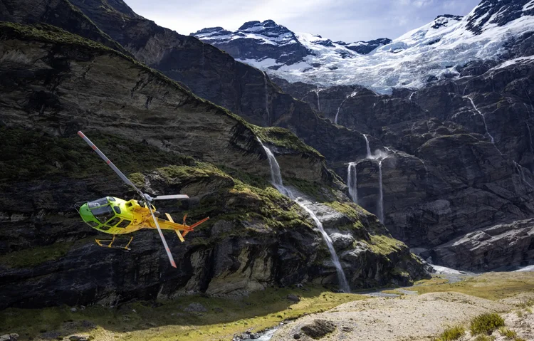 A helicopter flying near steep mountain slopes with waterfalls, snow-capped peaks, and lush green vegetation.