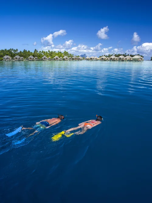 Two people snorkeling in clear blue water near overwater bungalows on a tropical island.