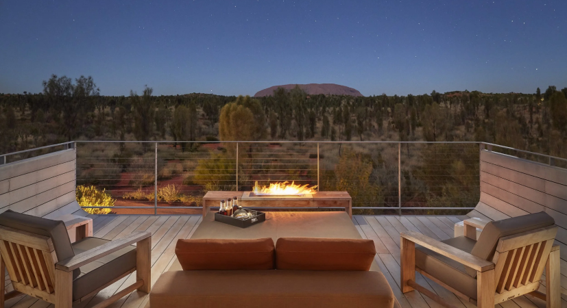 Outdoor deck at night with wooden furniture, a fire pit, and a view of a forest and flat mountain under a starry sky.