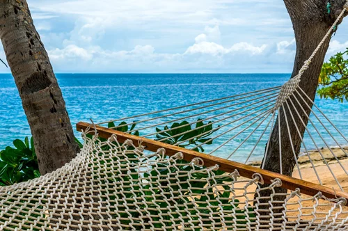 A hammock tied between two trees on a beach with the ocean in the background and partly cloudy sky.