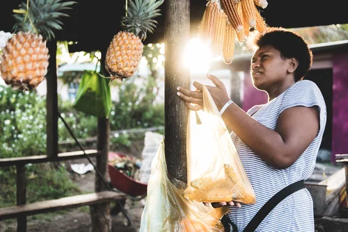 A woman standing at a street market stall hanging bunches of corn and pineapples, holding an orange plastic bag with food, with the sun shining behind her.