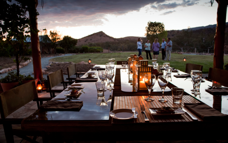 Outdoor dinner table set with glassware, plates, and candles at sunset, with a group of people standing and talking in the background on a grassy lawn surrounded by trees and mountains.