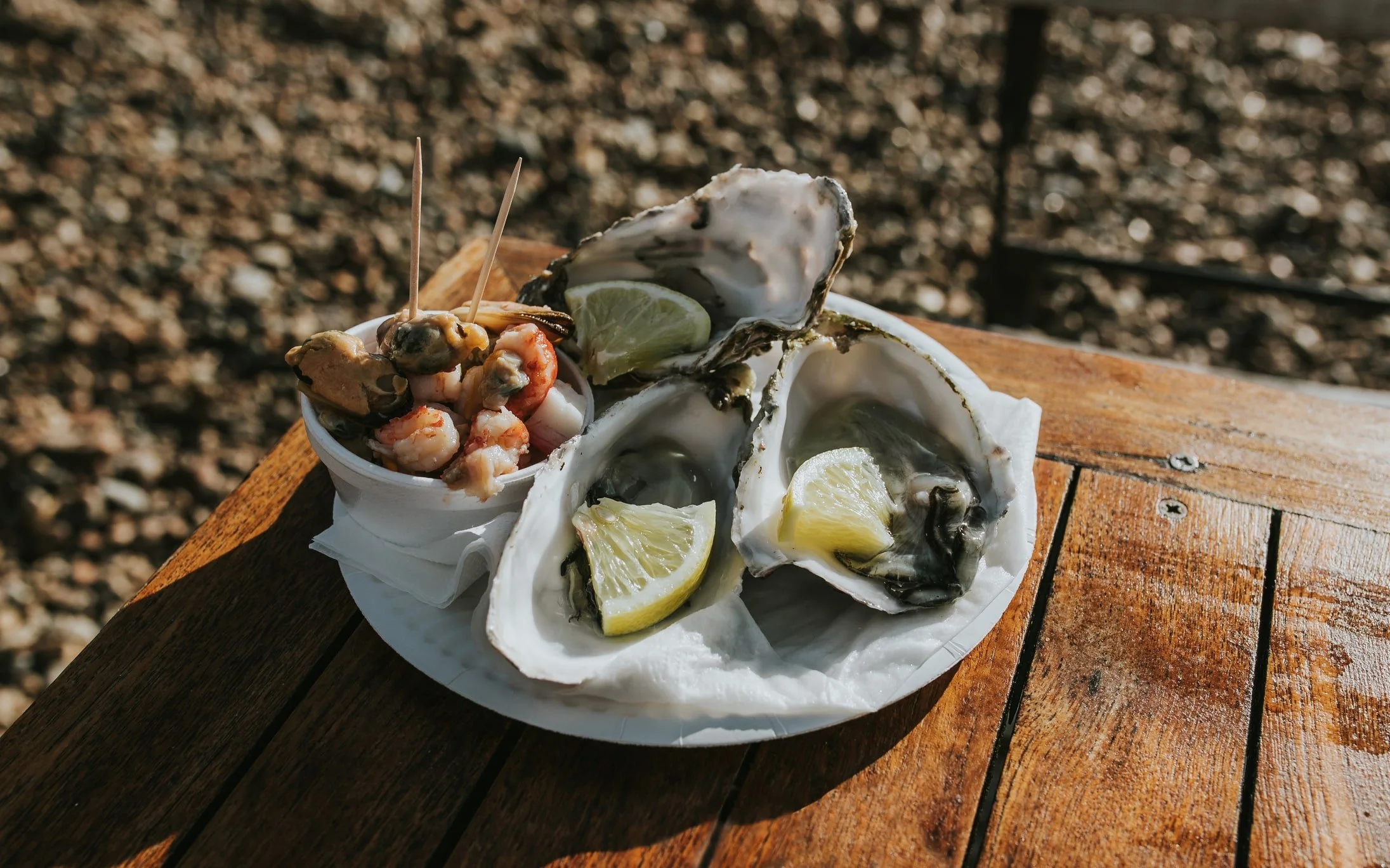 Zodiac oysters with lemon wedges served on a paper plate, accompanied by a small cup of seafood cocktail on a wooden table.