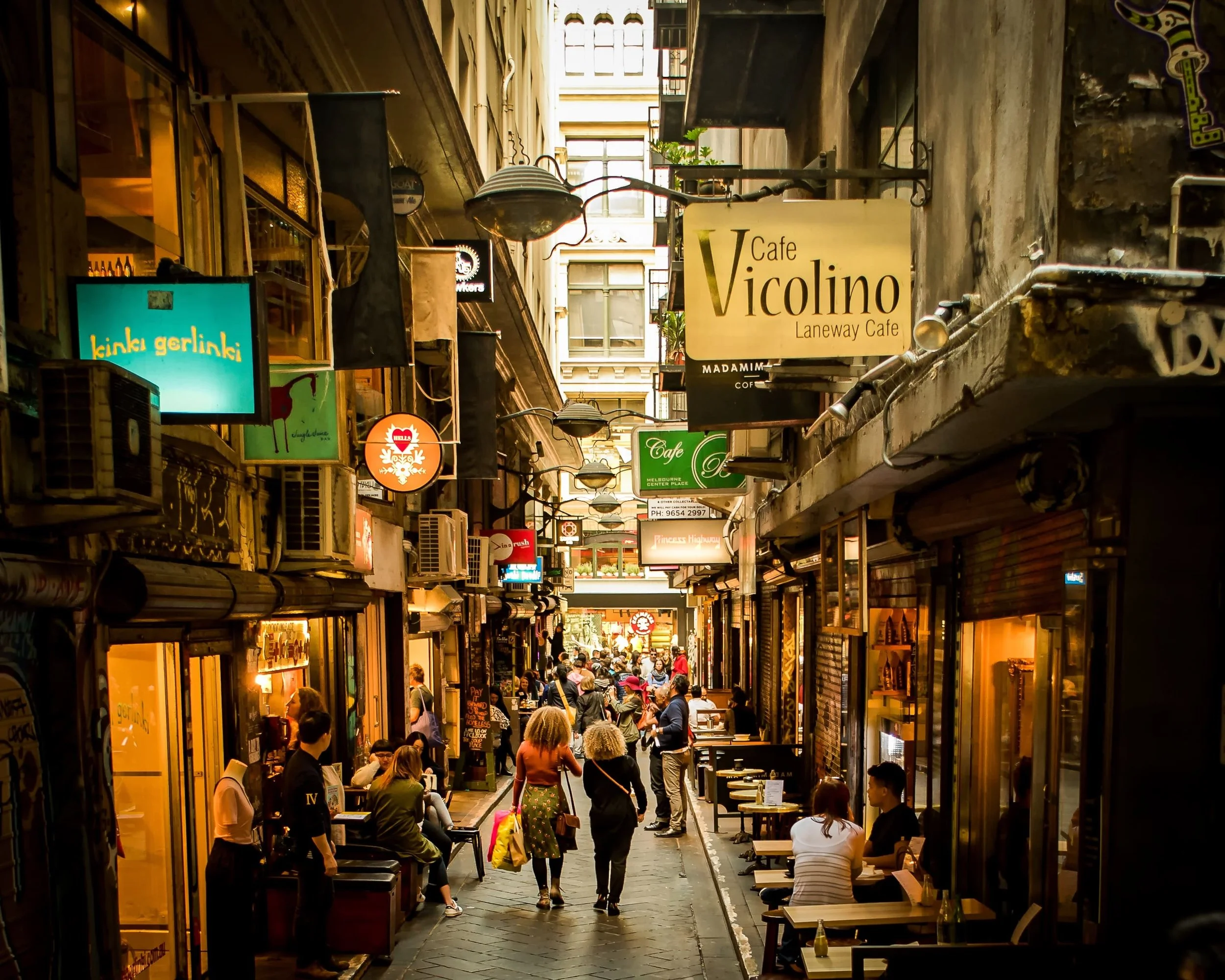 A bustling alleyway with outdoor seating at cafes, filled with people walking, sitting, and socializing during daytime.